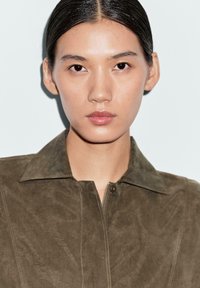Giovane con capelli scuri e lisci, che indossa una giacca in suede verde oliva con zip, rivolto in avanti con un'espressione neutra su uno sfondo semplice.