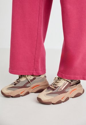 Masywne beżowo-brązowe sneakersy z pomarańczowymi akcentami noszone z różowymi spodniami o szerokich nogawkach na białej podłodze.