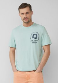 Lichtgroene katoenen t-shirt met korte mouwen, voorzien van een donkerblauw grafisch ontwerp en tekst aan de voorkant. Gemaakt met lichtoranje broeken.