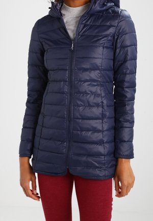 Femme portant une veste matelassée bleu marine zippée avec une capuche et un pantalon rouge, debout devant un fond blanc uni.