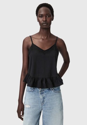 Frau trägt ein schwarzes ärmelloses Top mit Spitzenbesatz und gerafftem Saum, kombiniert mit hellblauen zerstörten Jeans, steht vor einem schlichten Hintergrund.