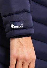 Marinblå vadderad jacka med quiltad design. Har elastiska muddar och en vit etikett med texten "By Superdry" på ärmen.