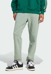 Pantaloni sportivi de culoare verde deschis, cu o textură netedă, croială conică și detalii pe cusăturile laterale, asortați cu adidași negri și albi.