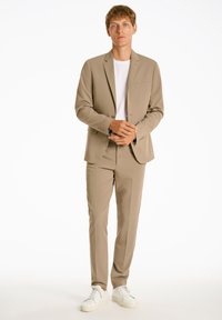 Beige Anzug mit einem Ein-Knopf-Blazer und passender schmal geschnittener Hose, kombiniert mit einem weißen T-Shirt und weißen Sneakern. Schlichte Designdetails.