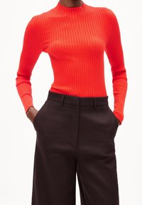 Person trägt ein leuchtend rotes, geripptes Langarmshirt, das in hochtaillierte, schwarze, weite Hosen gesteckt ist, mit den Händen in den Taschen.