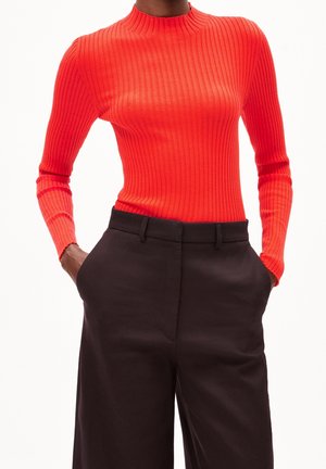 Persona vestida con una camiseta de manga larga acanalada de un rojo brillante, metida en unos pantalones anchos de tiro alto, negros, con las manos en los bolsillos.