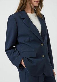 Námorně modrý přizpůsobený blazer s texturovanou tkaninou, jeden řádek knoflíků, špičaté klopy a dvě přední kapsy. Kovové knoflíkové detaily.