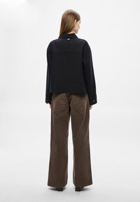 Casaco preto curto com design estruturado, apresentando gola e punhos com botões, combinado com calças largas castanhas com bolsos traseiros.