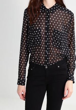 Frau trägt eine schwarze transparente gepunktete Bluse mit Knopfleiste vorne und Brusttaschen, kombiniert mit hoch taillierter schwarzer Hose.