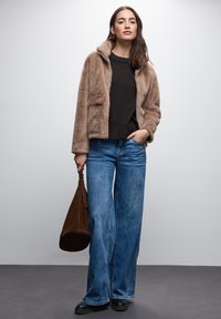 Beige Kunstfelljacke mit Kragen, schwarzer Pullover, weite blaue Jeans und braune Wildlederhandtasche. Schwarze Schuhe runden das Outfit ab.