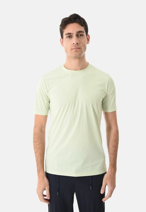 Giovane uomo di fronte che indossa una maglietta a maniche corte verde chiaro e pantaloni scuri su uno sfondo bianco uniforme.