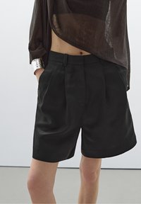 Personne portant un short noir taille haute sur mesure et un haut transparent marron foncé, main dans la poche, avec un large bracelet manchette argenté au poignet.