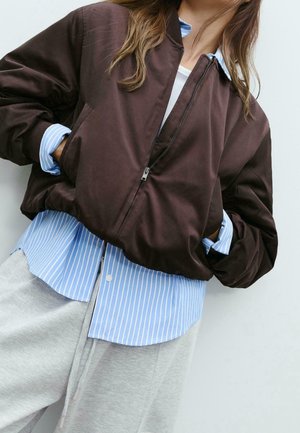 Personne portant une veste à fermeture éclair marron foncé par-dessus une chemise à rayures bleues et blanches avec boutons, et un pantalon de jogging gris clair, mains dans les poches de la veste.