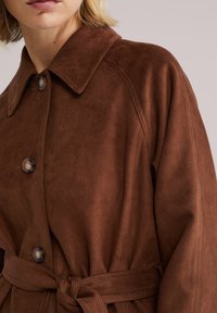 Palton trenci din piele întoarsă maro, cu un guler mare, nasturi și talie cu curea. Prezintă o textură netedă și detalii contrastante de cusături.