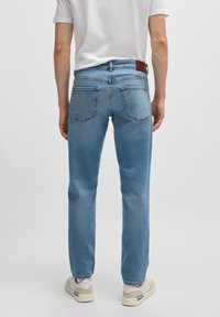 Una persona vestida con una camiseta blanca y jeans de color azul claro se encuentra de espaldas, mostrando los bolsillos traseros de los jeans y una etiqueta de cuero de marca.