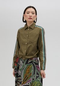 Olive green button-up shirt with white and green striped sleeves, featuring a chest pocket. Paired with a colorful, patterned skirt.