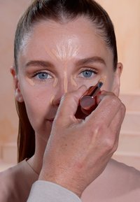 Applicazione del trucco con un stick correttore in crema. Un modello con occhi blu e pelle chiara ha il prodotto applicato in un motivo sul viso.