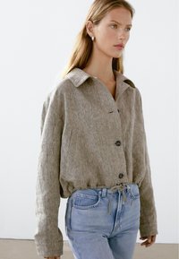 Vrouw in een losse, knoopbeige linnen jas met een trekkoord in de taille, gecombineerd met lichtblauwe, hoge spijkerbroeken, staand in de zij.