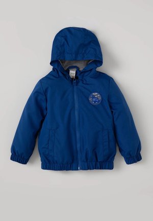 Giacca a vento blu con cappuccio, polsini elasticizzati e orlo regolabile. Presenta una toppa circolare con il testo "ROAR" e dei puntini.