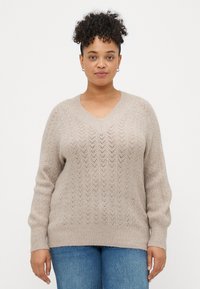 Beige strikket genser med V-hals, med et chevronmønster og ribbestrikkede mansjetter. Myk tekstur, avslappet passform og noe oversized design.