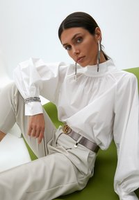 Biela blúzka s dlhými rukávmi a nariaseným výstrihom, skombinovaná s svetlo béžovými nohavicami a strieborným pásom. Obsahuje veľké kovové náušnice.