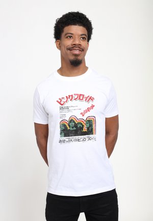 Mann mit kurzen lockigen Haaren und Ziegenbart, der ein weißes Pink Floyd Band Grafik-T-Shirt trägt, steht mit den Händen hinter dem Rücken, schaut nach rechts und lächelt.