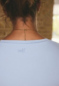 Top azul claro con canalé y escote ancho, adornado con un detalle bordado de "self." en la parte posterior, combinado con un delicado collar de cadena dorada.