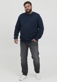 Suéter de punto azul marino con cuello redondo, puños y dobladillo acanalados, combinado con jeans gris medio y zapatillas blancas. El modelo está de pie contra un fondo liso.