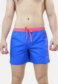 Pantaloni da nuoto blu con vita e cordino rosa. Presentano il logo 'SUNDEK' in rosa sul lato. Realizzati in un materiale leggero e morbido.