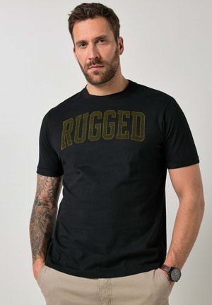 Bärtiger Mann mit tätowiertem linken Arm, der ein schwarzes T-Shirt mit dem Schriftzug "RUGGED" und beige Hosen trägt, Hände in den Taschen, neutraler Hintergrund.