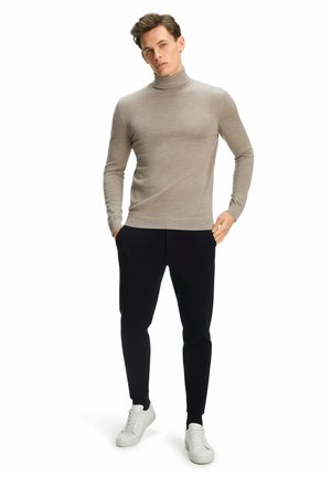 Beige coltrui van lichtgewicht stof, gecombineerd met donkere broeken en witte sneakers. Model staat met handen in de zakken, neutrale achtergrond.