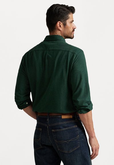 Mann som står med ryggen til iført en mørkegrønn langermet skjorte med oppbrettede ermer og mørkeblå jeans med et brunt belte.