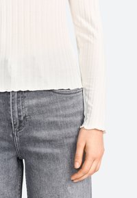 Top bianco a maniche lunghe con orlo ondulato, abbinato a jeans grigi a vita alta. Texture liscia e design aderente.