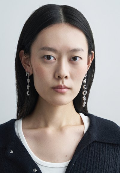 Silberne Ohrringe mit hängenden Buchstaben, die "MARC JACOBS" buchstabieren, sind an einem Modell zu sehen, das einen dunklen Pullover über einem weißen Oberteil trägt.