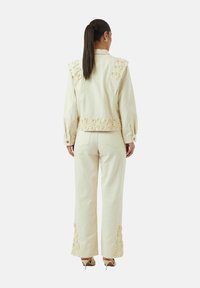 Conjunto de dos piezas color crema que incluye una chaqueta con bordados florales en los hombros y el dobladillo, y pantalones a juego con detalles decorativos en los tobillos.