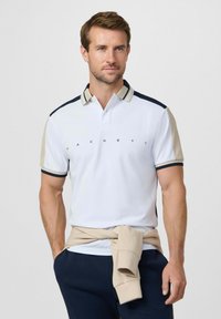 Man met een wit poloshirt met beige en navy strepen, een beige sweatshirt om de taille gestrikt, staand met de handen in de zakken van navybroek.