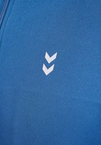 Blå textil med en slät yta har en vit chevronlogotyp bestående av två nedåtriktade former på den övre vänstra sidan.