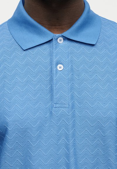 Κοντινό πλάνο ενός ατόμου με σκούρο τόνο επιδερμίδας που φοράει ανοιχτό μπλε πικέ πόλο πουκάμισο με μυτερό γιακά και δύο λευκά κουμπιά κουμπωμένα.