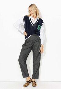 Gilet en tricot marine avec des finitions blanches et la lettre "B" verte, porté sur une chemise blanche, associé à un pantalon à carreaux gris et des chaussures imprimé léopard.