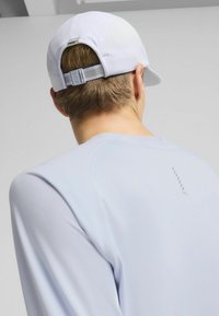 Jasnoniebieska, sportowa koszulka z długim rękawem o gładkiej fakturze, zaokrąglonych ramionach oraz odblaskowym logo. Biała czapka z siateczkowymi akcentami.