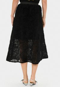 Fustă midi neagră din dantelă texturată cu un model floral, având o croială dreaptă și un overlay transparent, asortată cu sandale cu toc argintii.