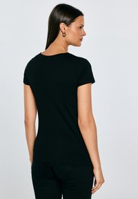 Hosszú sötét hajú nő, egy egyszerű fekete rövid ujjú pólót és fekete nadrágot visel, hátulról látható világosszürke háttér előtt.