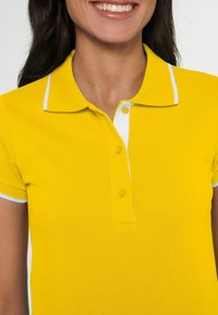 Geel polo shirt van textuurstof met een klassieke kraag, een knoopsluiting met drie knopen en witte bies op de kraag en mouwen.
