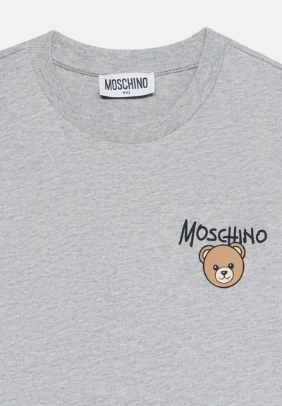 Ανοιχτό γκρι παιδικό μπλουζάκι με ετικέτα Moschino και μικρό γραφικό με πρόσωπο αρκουδάκι δίπλα στο όνομα της μάρκας Moschino μπροστά.