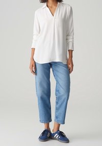 Haut blanc à manches longues avec un col en V et un ourlet arrondi, associé à un jean bleu clair ample et des chaussures de sport bleu marine.