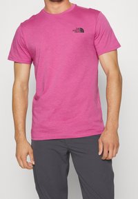 Růžové bavlněné tričko s kulatým výstřihem a krátkými rukávy, s černým logem na levé straně hrudi. Hladká textura, volný střih.