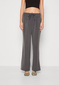 Pantalons larges gris avec une taille élastique et un cordon de serrage, fabriqués à partir d'un tissu lisse. Ils sont associés à des sandales vertes à lanières.