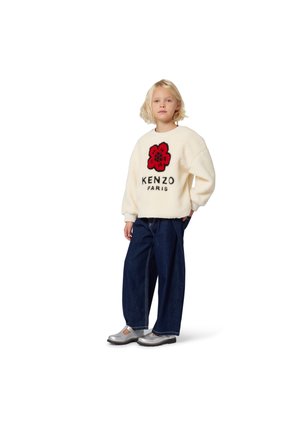 Kremowy polarowy sweter z czerwonym kwiatowym wzorem i czarnym napisem "KENZO PARIS". Zestawiony z ciemnoniebieskimi szerokimi spodniami dżinsowymi i srebrnymi butami.
