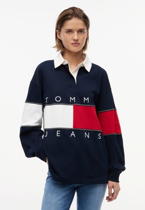 Jeune personne portant un maillot de rugby manches longues bleu marine, blanc et rouge avec le texte "TOMMY JEANS", debout devant un fond clair uni.