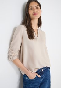 Blouse légère beige clair avec un col en V et des manches longues retroussées, associée à un jean bleu foncé. Texture lisse et coupe décontractée.